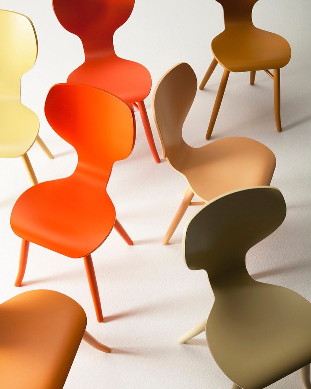 Several modern chairs in varying orange, yellow, and brown hues are arranged on a bright white surface, creating a colorful display.