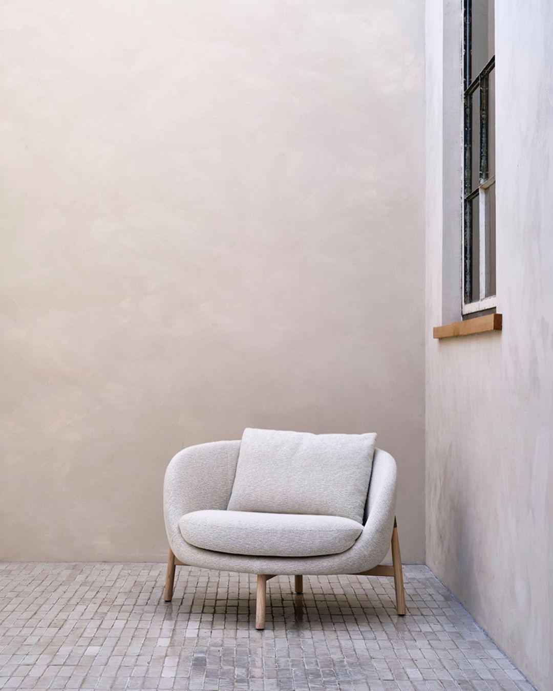 Indoor shot of a modern, light grey armchair with a cushion, set on a tiled floor against a neutral wall with window.