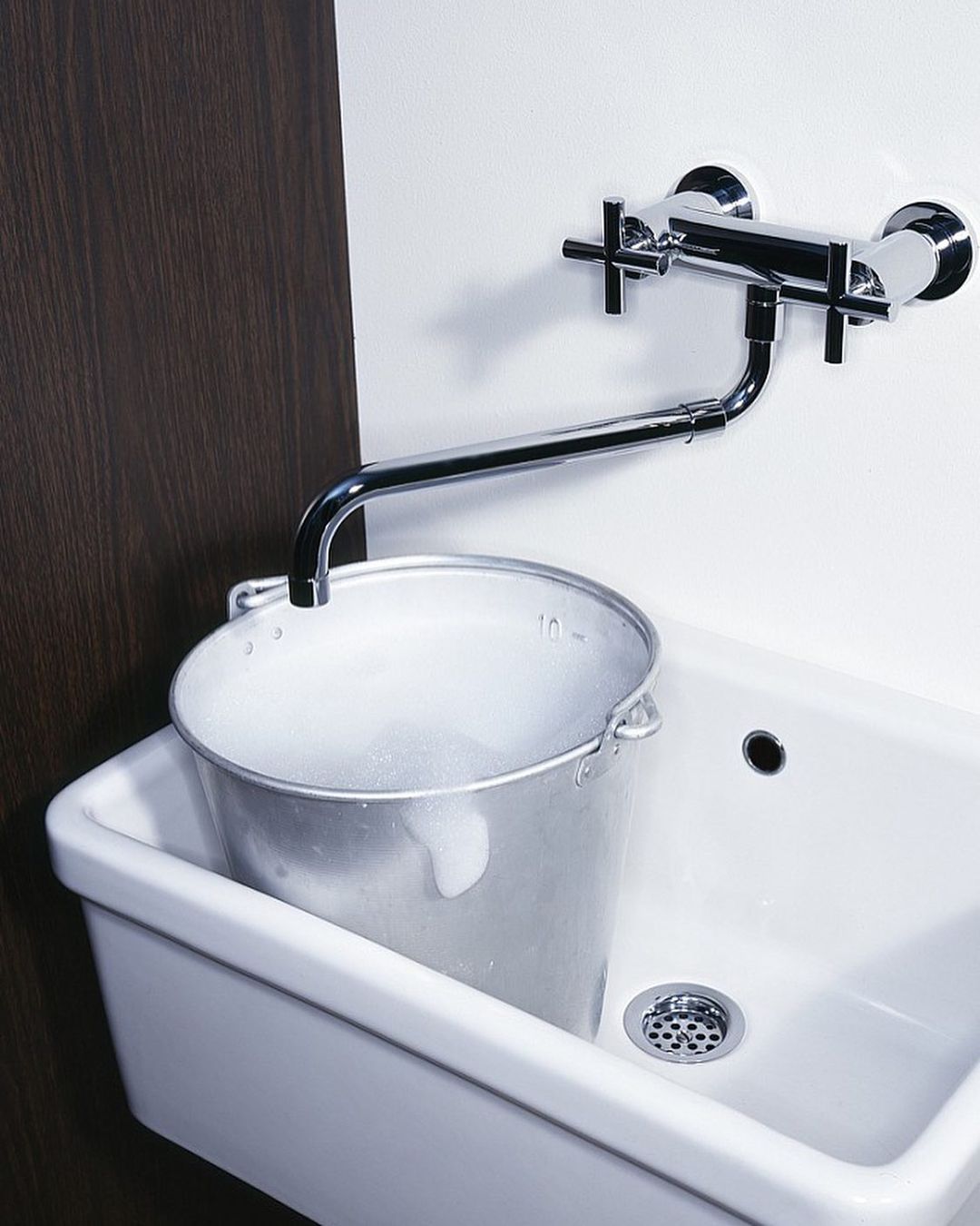 Chrome faucet dispenses water into a galvanized metal bucket, set inside a white porcelain sink with a dark brown wall background.