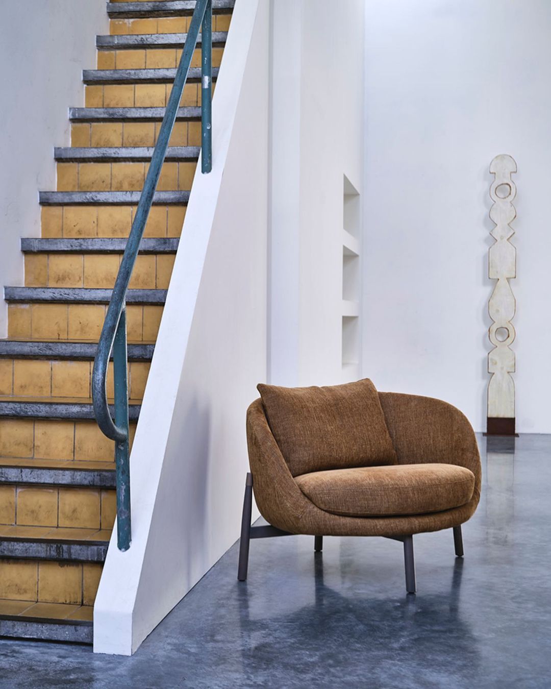 Upholstered armchair with pillow on a concrete floor, with a staircase and geometric sculpture in the background, in a bright interior space.