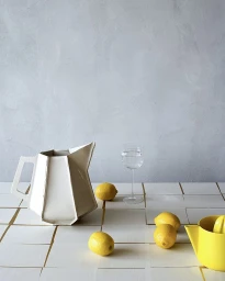 White faceted ceramic pitcher, yellow lemons, a glass of water, and a yellow juicer on a tiled tabletop.