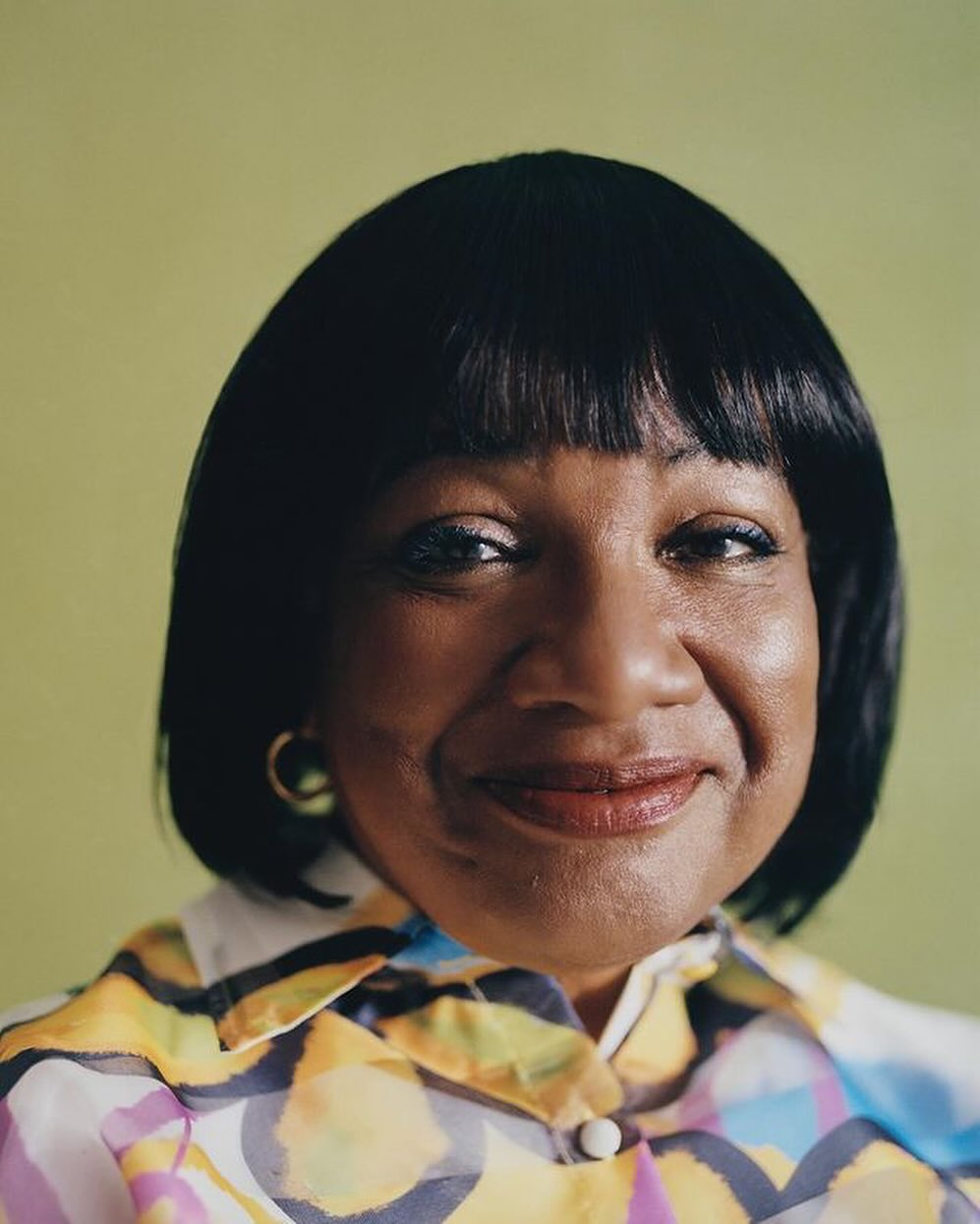 Close-up portrait of an adult woman with dark hair and a colorful patterned shirt, smiling in front of a light green background.