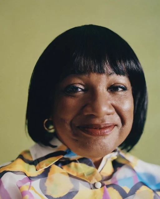 Close-up portrait of an adult woman with dark hair and a colorful patterned shirt, smiling in front of a light green background.