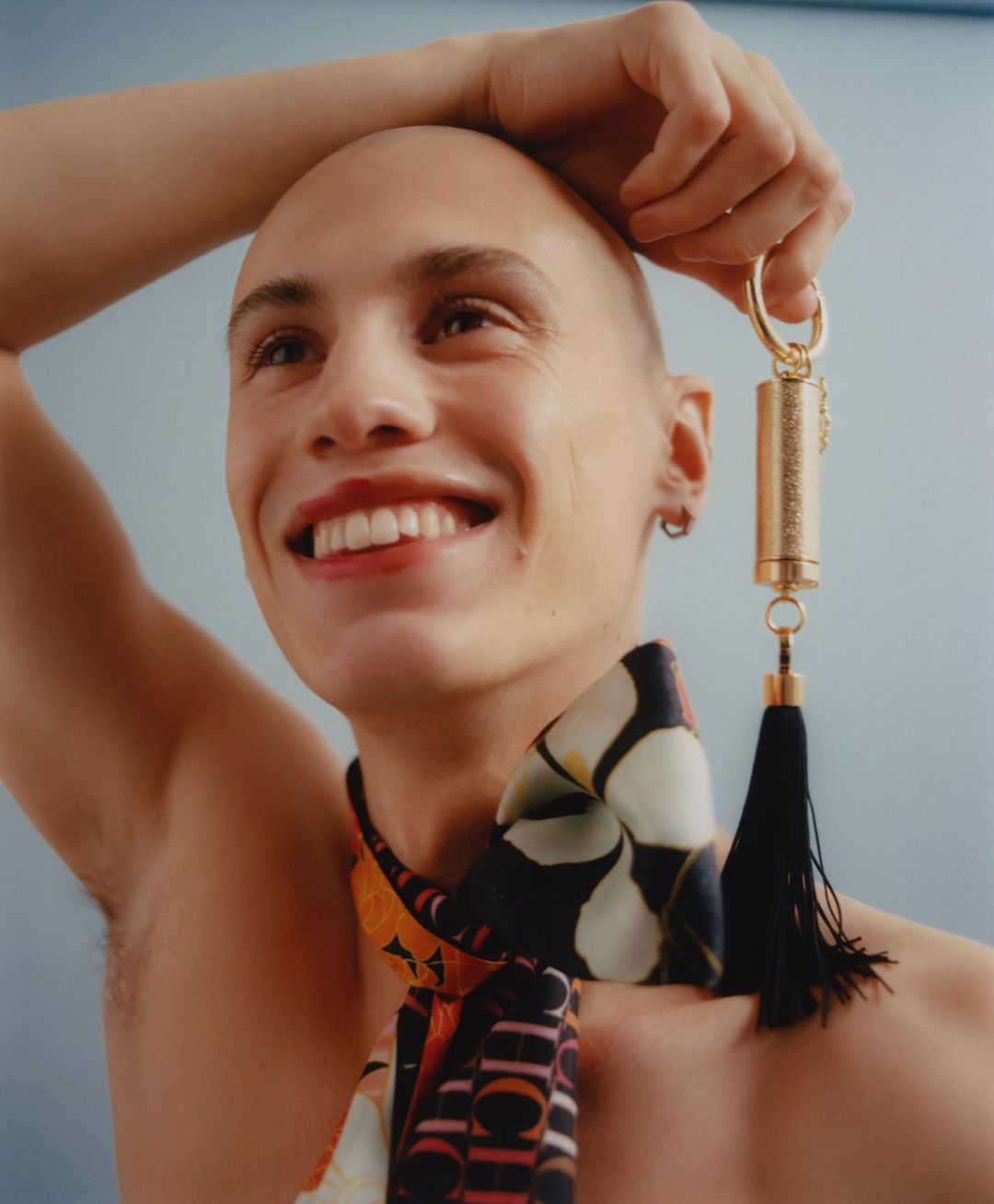 Smiling, bare-chested adult with bald head holds gold cylinder object with tassel and a colourful scarf, set against a blue background.