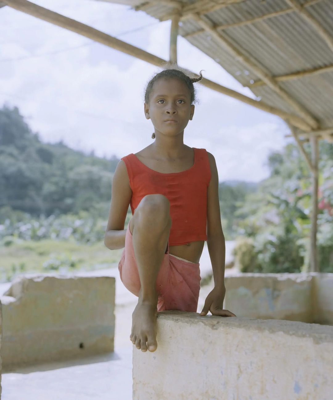 A young person sits on a ledge wearing a red sleeveless top and pink skirt, with a dense green forest visible in the background.