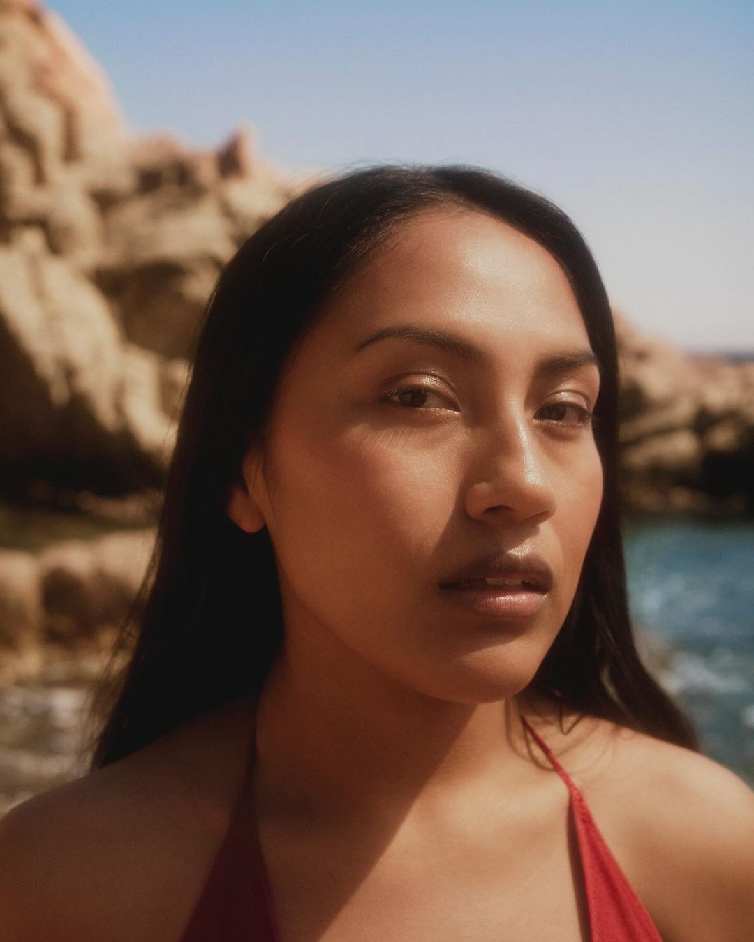 Close-up portrait of a woman with long dark hair wearing a red top against a rocky coast and sea by Silvana Trevale.