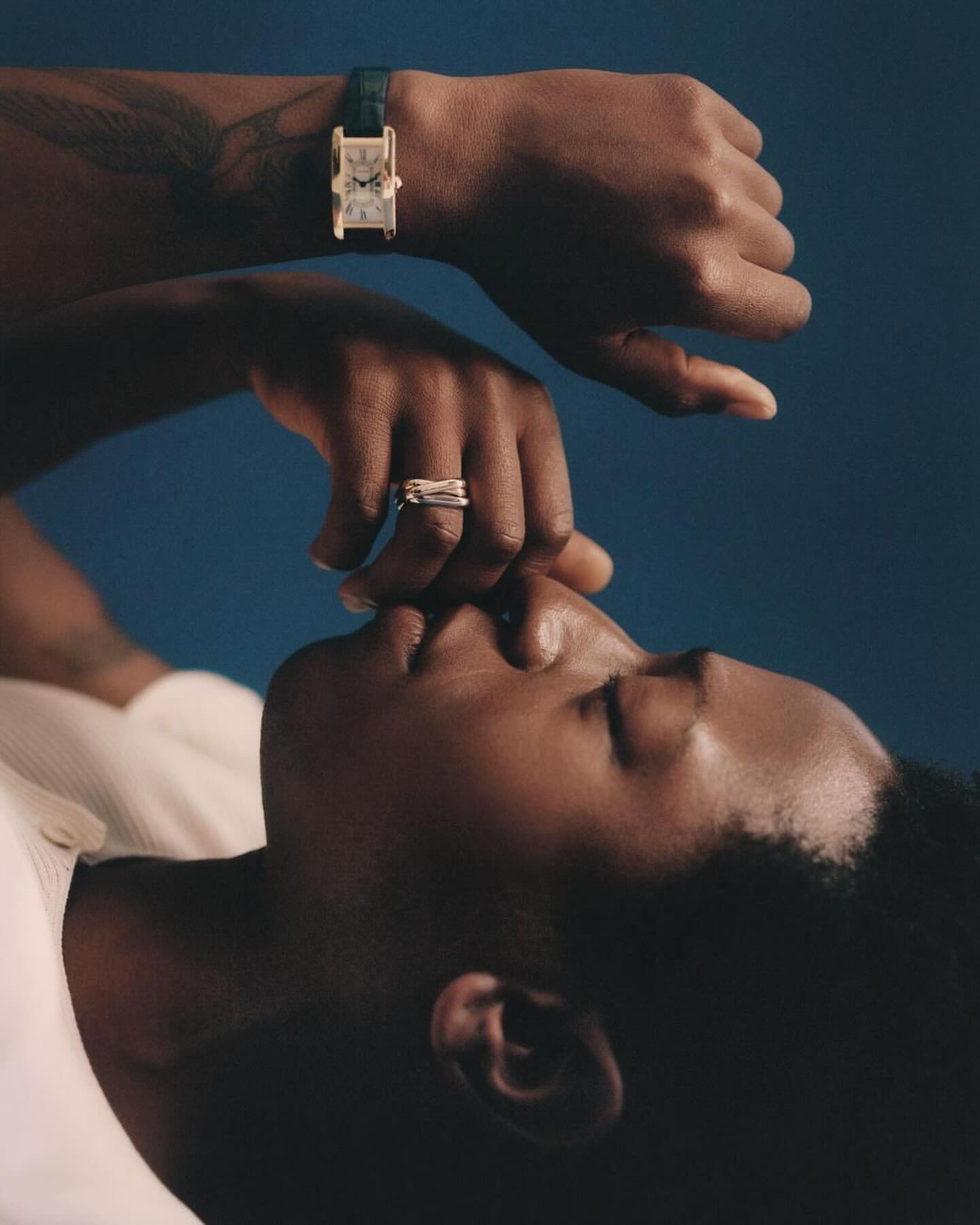 Portrait of a Black person lying down, wearing a wristwatch and rings, against a dark blue backdrop in soft, diffused lighting.