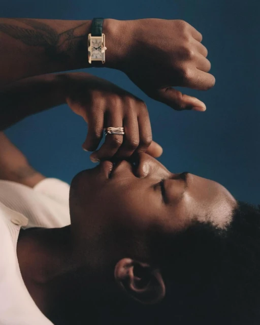 Portrait of a Black person lying down, wearing a wristwatch and rings, against a dark blue backdrop in soft, diffused lighting.