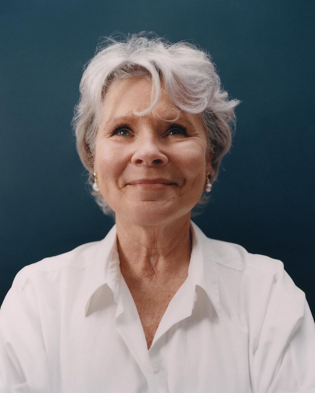 Close-up portrait of an elderly woman with gray hair wearing a white collared shirt against a dark teal background in eye-level shot.