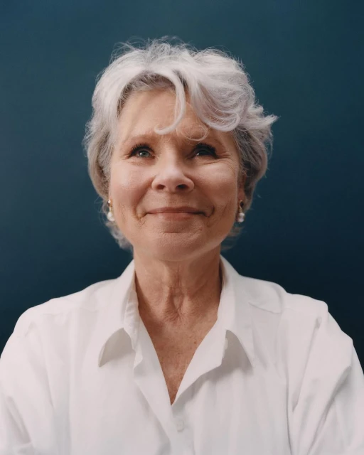 Close-up portrait of an elderly woman with gray hair wearing a white collared shirt against a dark teal background in eye-level shot.