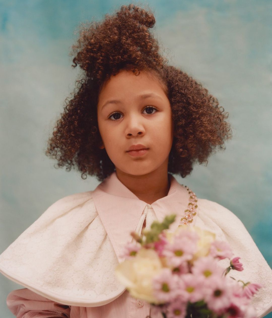 Portrait of a child with curly hair holding a bouquet of flowers against a blue background, wearing a cape and collared shirt. 