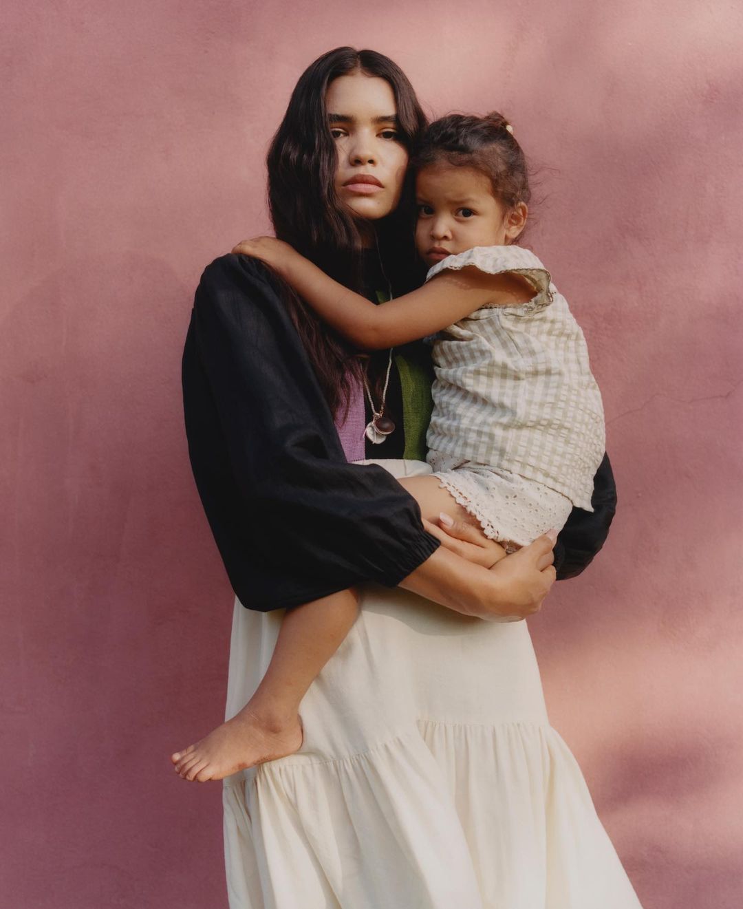 Portrait of a woman holding a young child in her arms against a textured pink plaster wall.