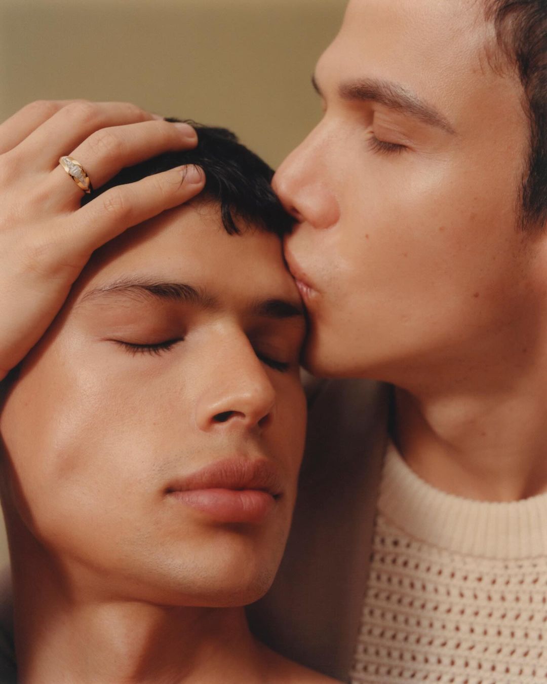 A close-up portrait captures a tender moment between two adult men. One man gently kisses the other on the forehead in a warm-toned setting.