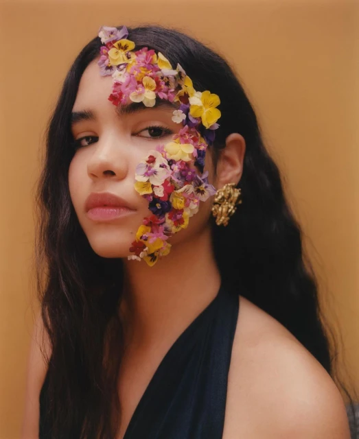 Close-up portrait of a woman with flowers on her face and wearing a black dress, set against a yellow background.