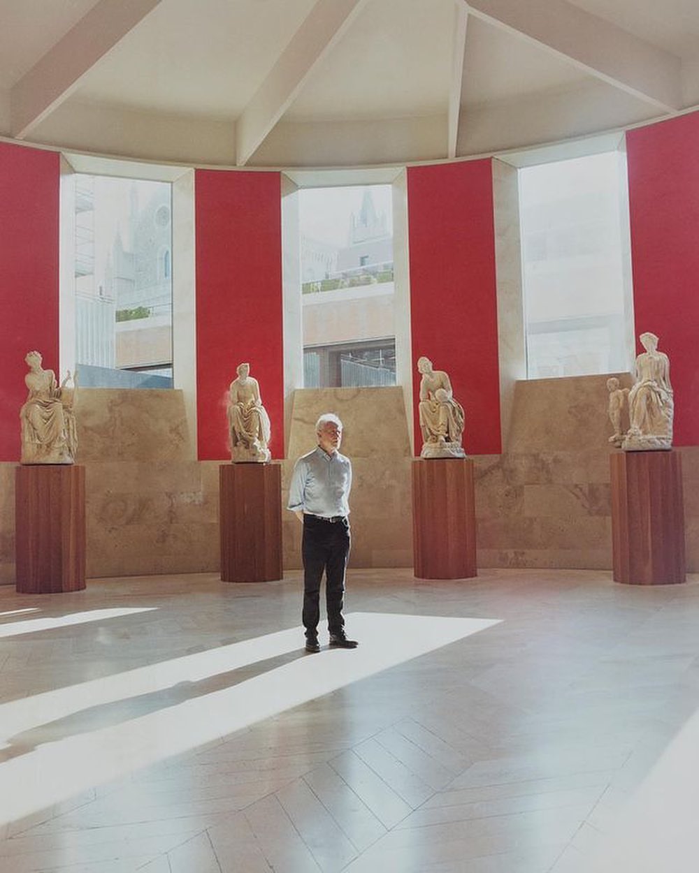 Mature man standing in a room with classical statues on pedestals and red accents near windows, with bright natural light and shadows.