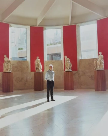 Mature man standing in a room with classical statues on pedestals and red accents near windows, with bright natural light and shadows.