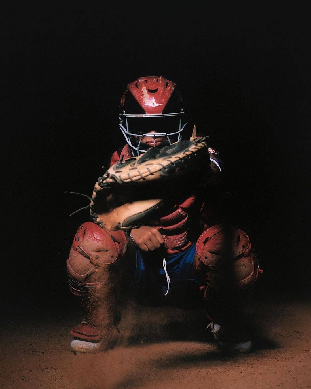 Baseball catcher in red gear crouches on a dirt field with dramatic lighting, ready to catch, creating a dynamic and sporty image.