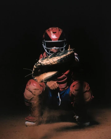 Baseball catcher in red gear crouches on a dirt field with dramatic lighting, ready to catch, creating a dynamic and sporty image.