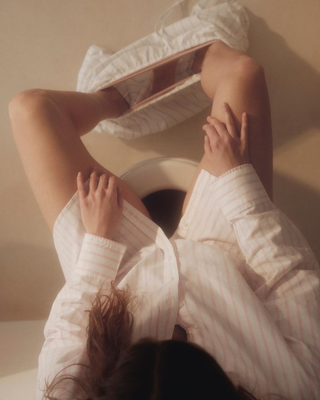 Woman in a striped shirt lies upside down on the floor with legs bent in front of a toilet, holding her knees with both hands.