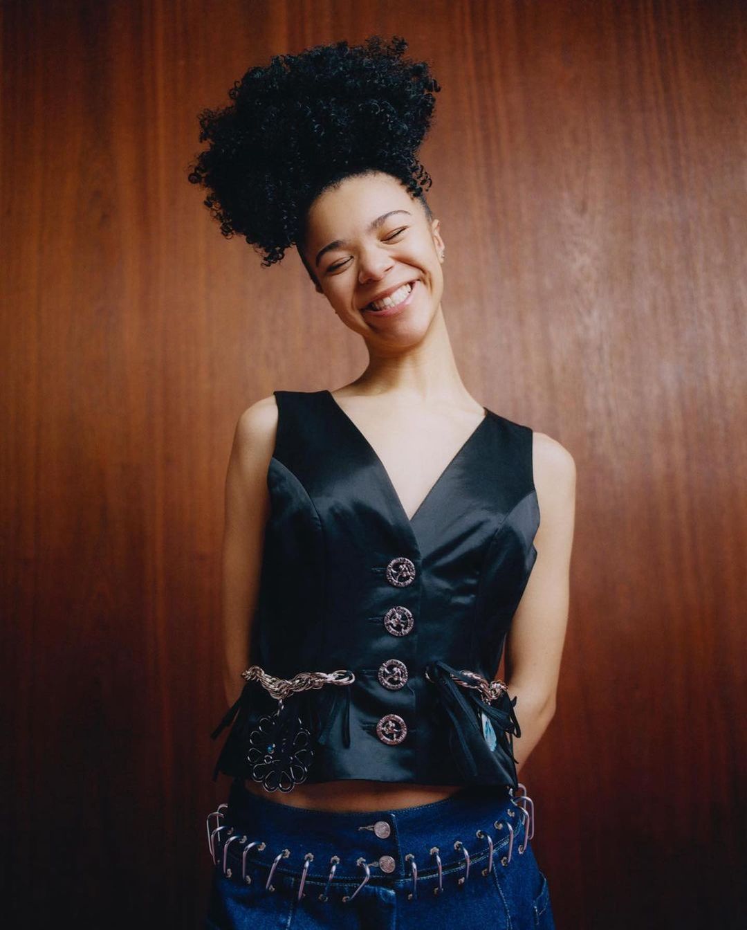 Young woman with an afro smiles in a close-up studio shot. She's wearing a black vest and blue jeans with a wood paneled background.