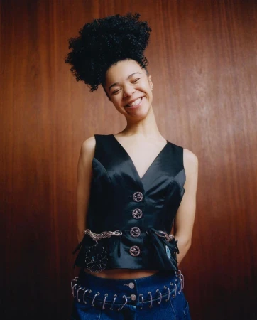 Young woman with an afro smiles in a close-up studio shot. She's wearing a black vest and blue jeans with a wood paneled background.