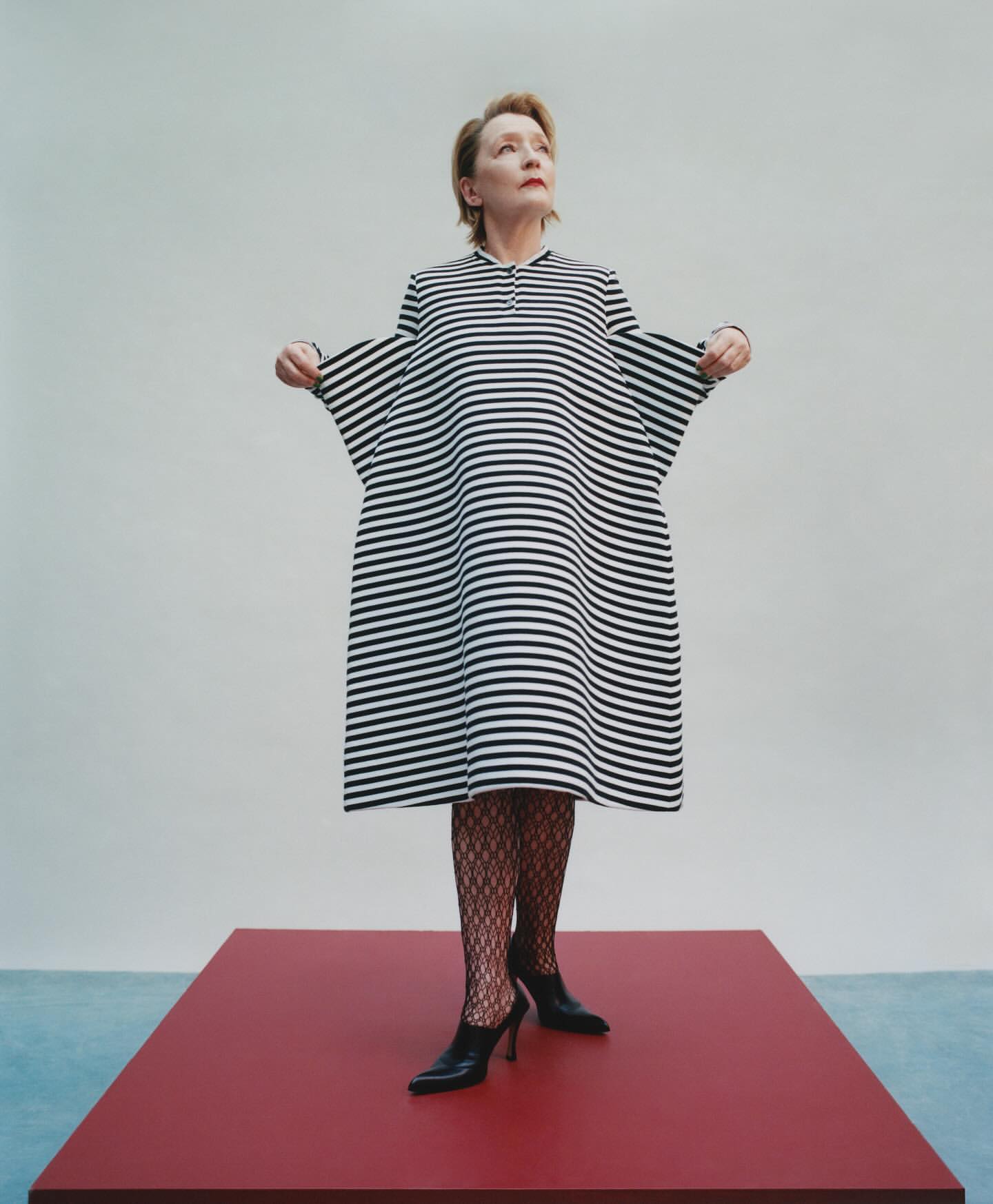 Elegant woman stands on red platform in a black and white striped dress and black heels, striking a fashionable pose in a photo studio.