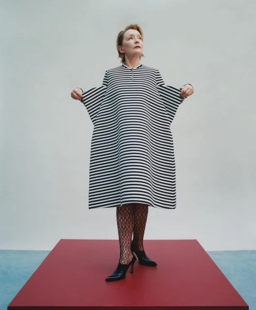 Elegant woman stands on red platform in a black and white striped dress and black heels, striking a fashionable pose in a photo studio.