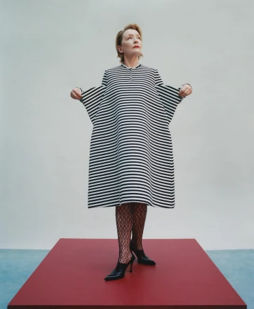 Elegant woman stands on red platform in a black and white striped dress and black heels, striking a fashionable pose in a photo studio.