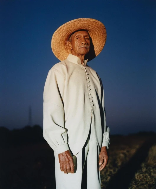 Portrait of an elderly Hispanic man wearing a wide-brimmed straw hat and cream-colored clothing against a clear blue sky at dusk.