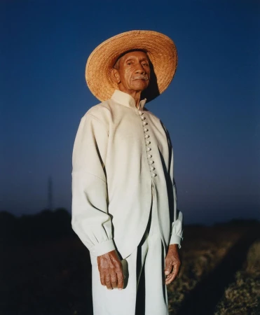 Portrait of an elderly Hispanic man wearing a wide-brimmed straw hat and cream-colored clothing against a clear blue sky at dusk.