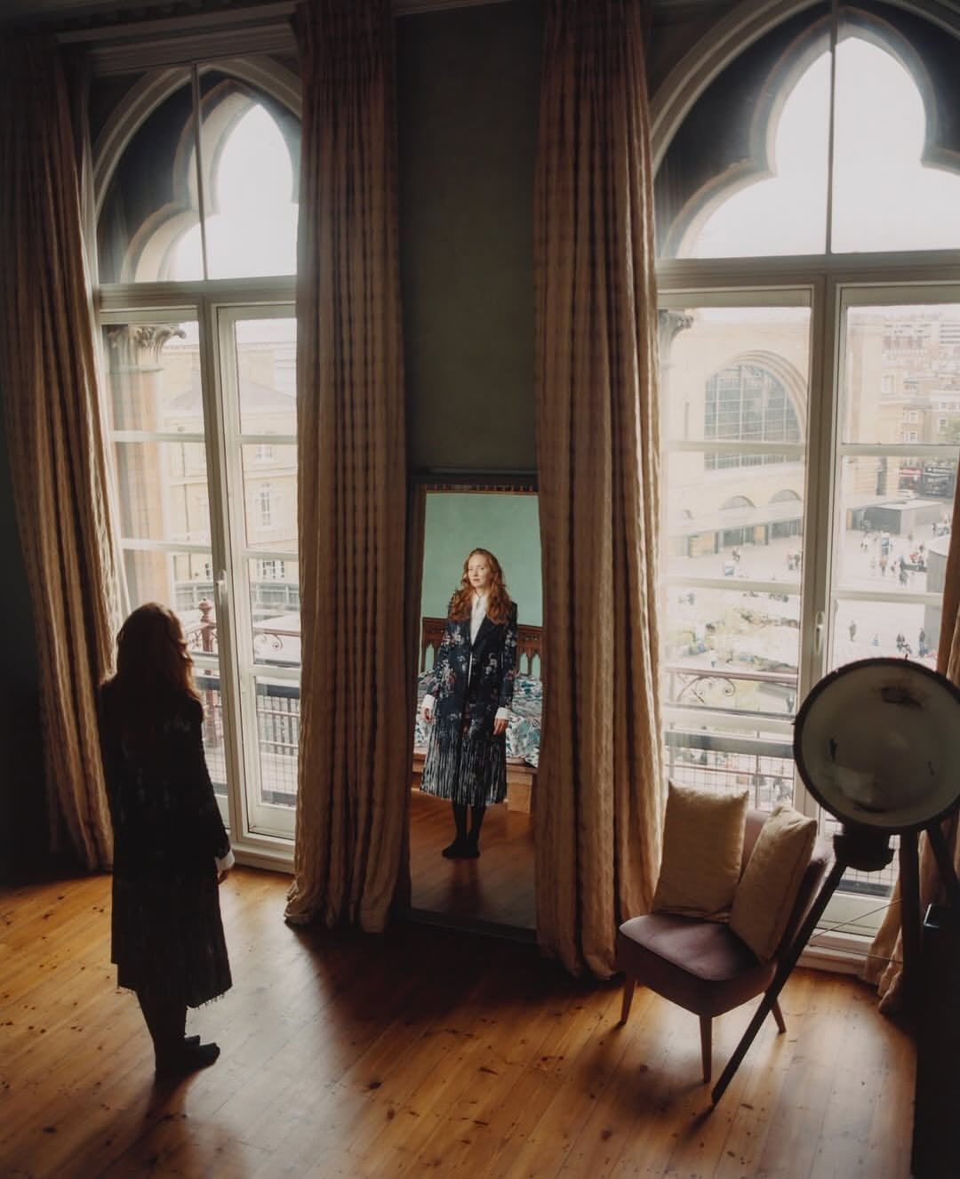 A woman looks in a mirror inside a room with large windows and drapes. Another woman stands in the room with a chair and a lamp.