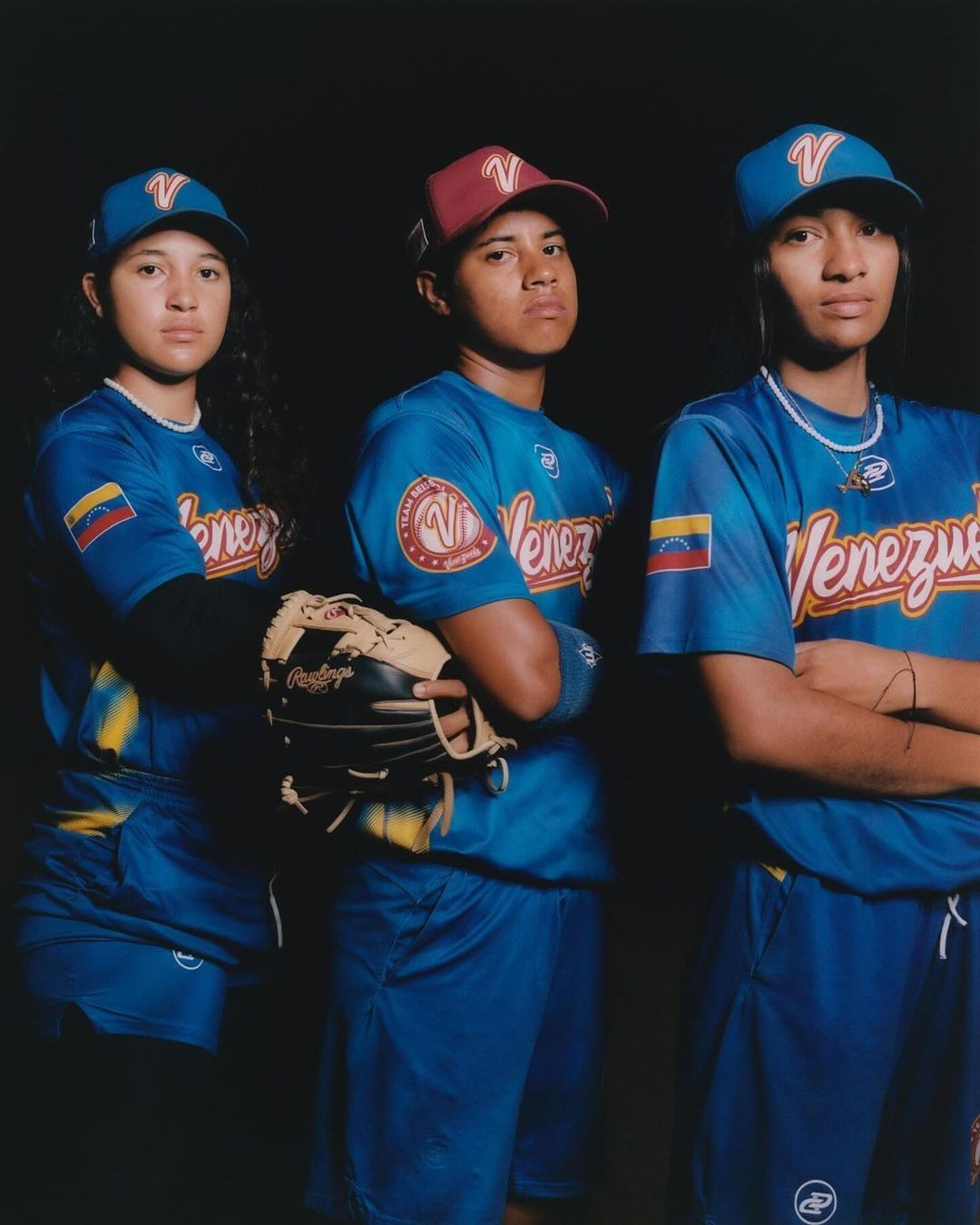 Three young female baseball players in blue Venezuela uniforms and hats pose indoors against a dark backdrop, one holding a baseball glove.