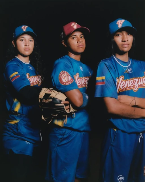 Three young female baseball players in blue Venezuela uniforms and hats pose indoors against a dark backdrop, one holding a baseball glove.
