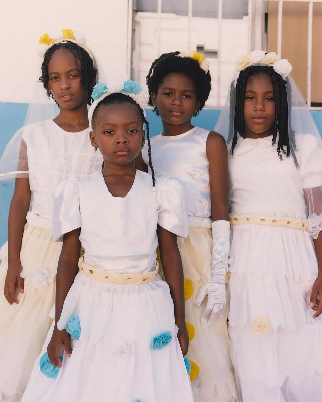 Four young Black girls in formal white dresses with flower crowns pose together against a light blue and white background for a portrait.
