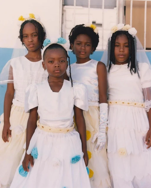 Four young Black girls in formal white dresses with flower crowns pose together against a light blue and white background for a portrait.