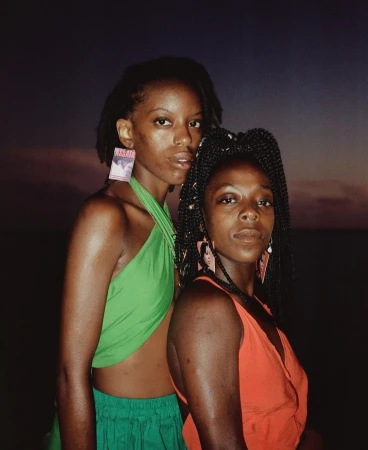 Two black women posing outdoors at dusk, wearing a green halter top and an orange halter top with statement earrings, captured by Silvana Trevale.