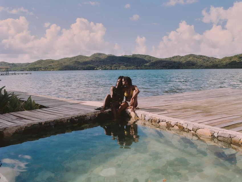Two Black adult females in swimwear sit near a pool overlooking a lake with islands under a bright sky with scattered clouds in daylight.