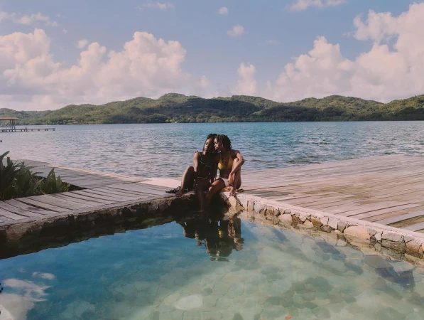Two Black adult females in swimwear sit near a pool overlooking a lake with islands under a bright sky with scattered clouds in daylight.