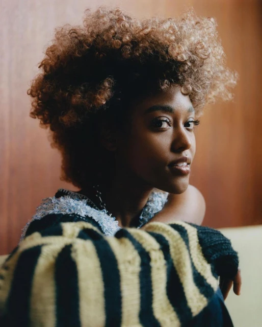 Close-up portrait of a young Black woman with curly hair, wearing a striped sweater, against a wood-paneled wall in a bright, soft studio.