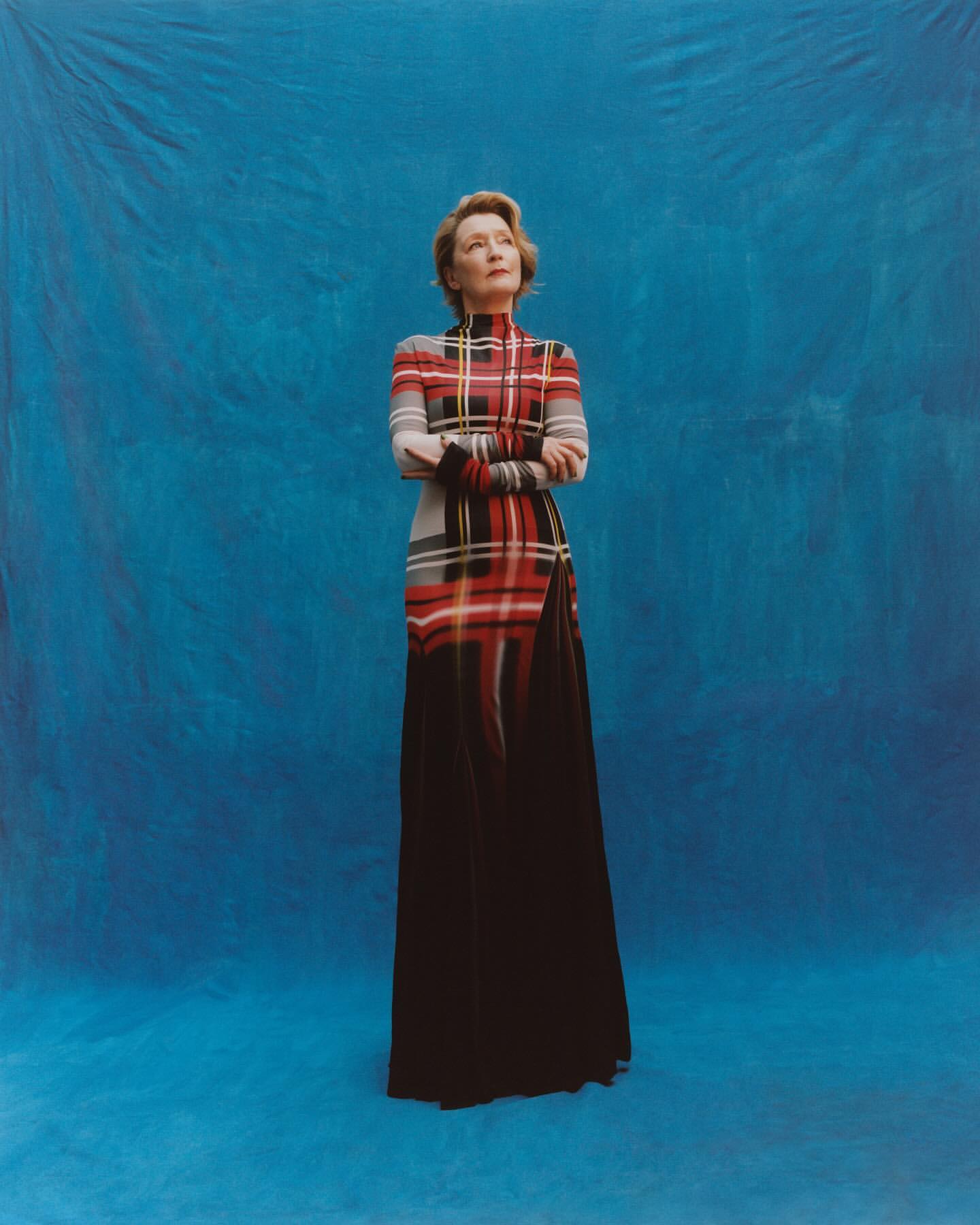 Elegant woman in a plaid dress with a high neckline poses in front of a blue backdrop, looking upward with her arms crossed.