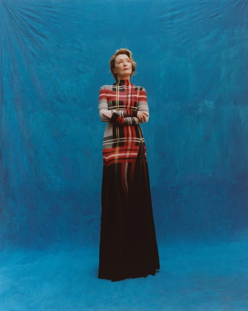 Elegant woman in a plaid dress with a high neckline poses in front of a blue backdrop, looking upward with her arms crossed.