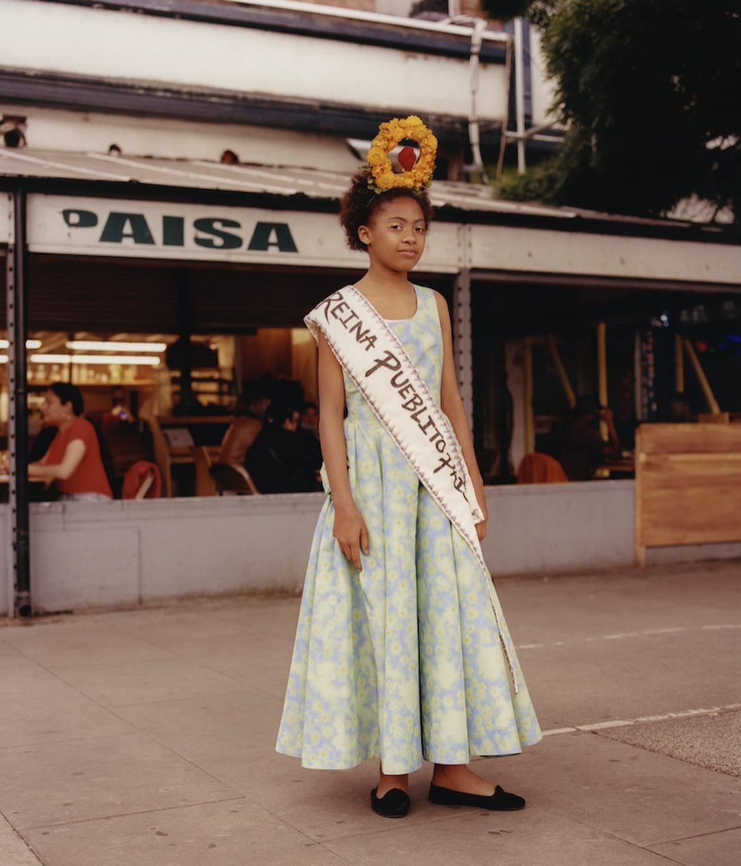 Young girl in floral dress with flower crown and sash stands on sidewalk in an urban environment with buildings in the background.