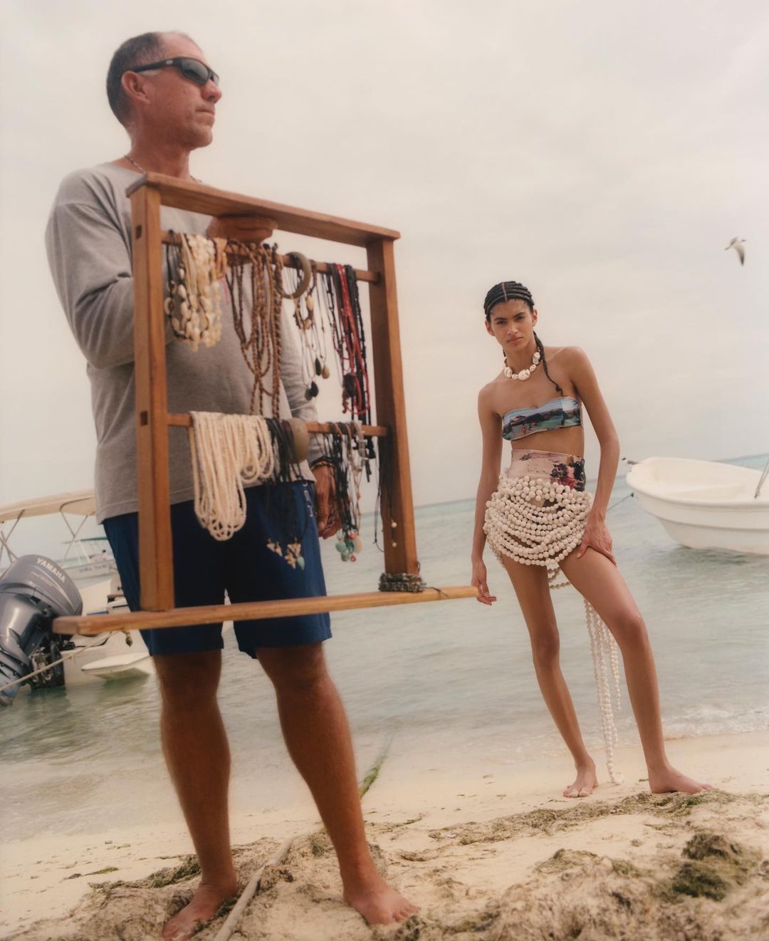 A man and a woman pose on a beach with jewelry on display, featuring a light blue sky, white sand, and a small white boat.