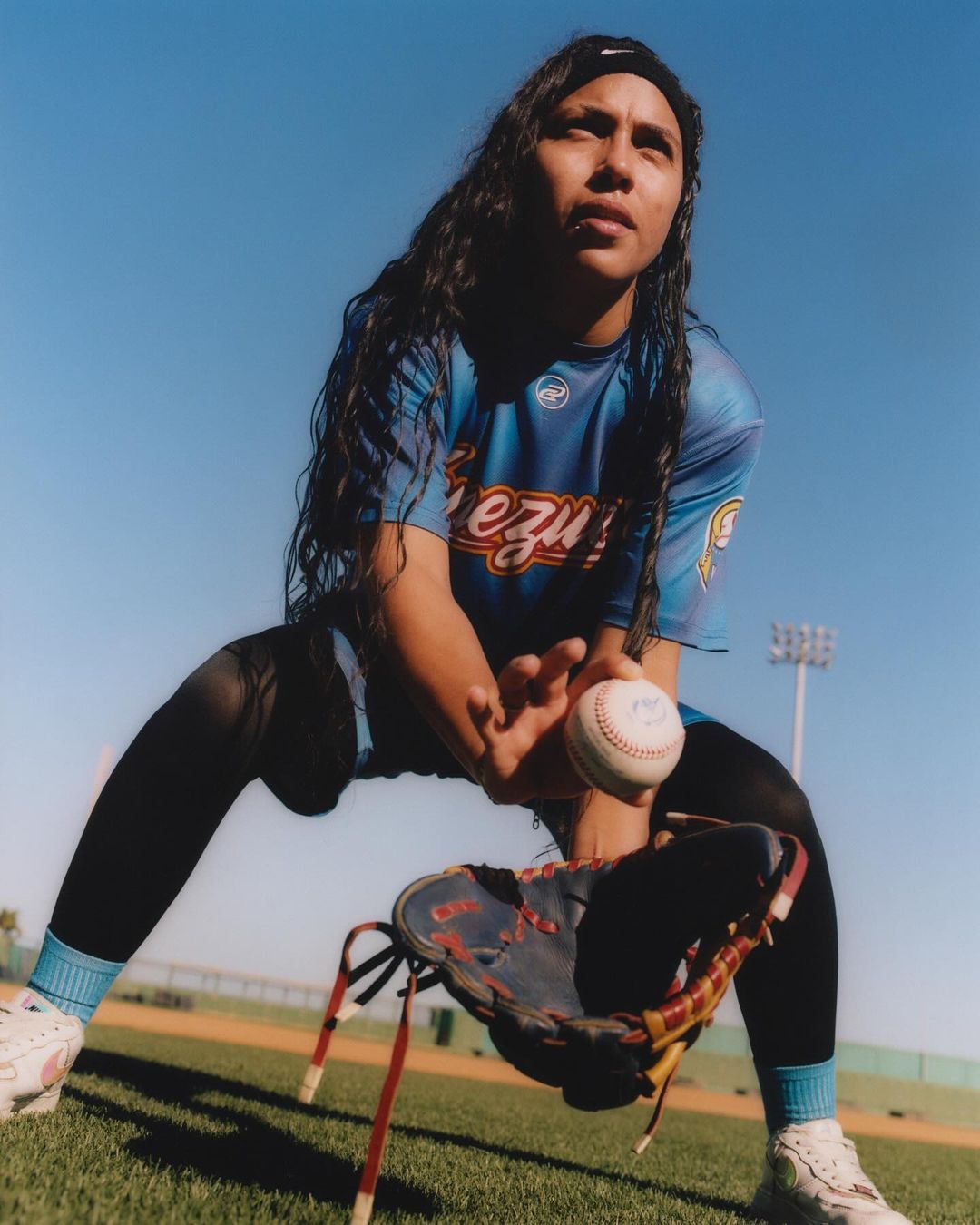Female baseball player in uniform fielding a baseball with mitt in a stadium on a sunny day with clear blue skies in an eye-level outdoor shot.