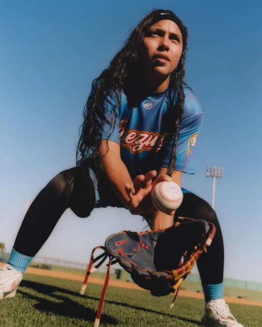 Female baseball player in uniform fielding a baseball with mitt in a stadium on a sunny day with clear blue skies in an eye-level outdoor shot.