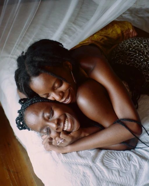 Two dark-skinned adult females are embracing and smiling on a bed covered with a white comforter, with a translucent canopy visible above.