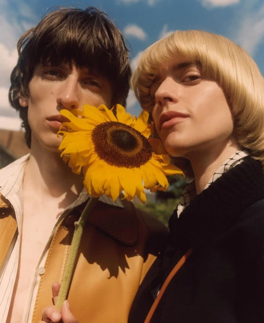 Two people pose with a yellow sunflower in an outdoor setting with a blue sky and clouds, creating a natural and engaging visual.