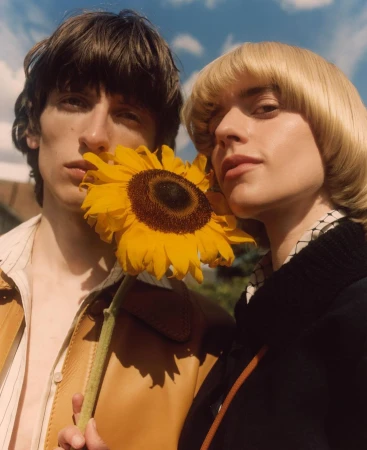 Two people pose with a yellow sunflower in an outdoor setting with a blue sky and clouds, creating a natural and engaging visual.