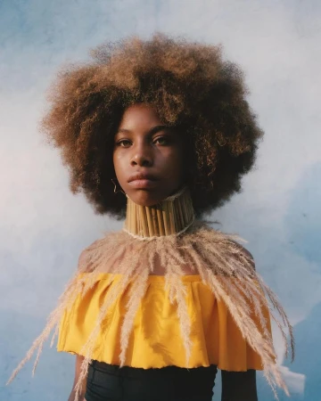 Portrait of a young Black girl with Afro hair and a decorative straw collar and shoulder piece against a neutral blue background.