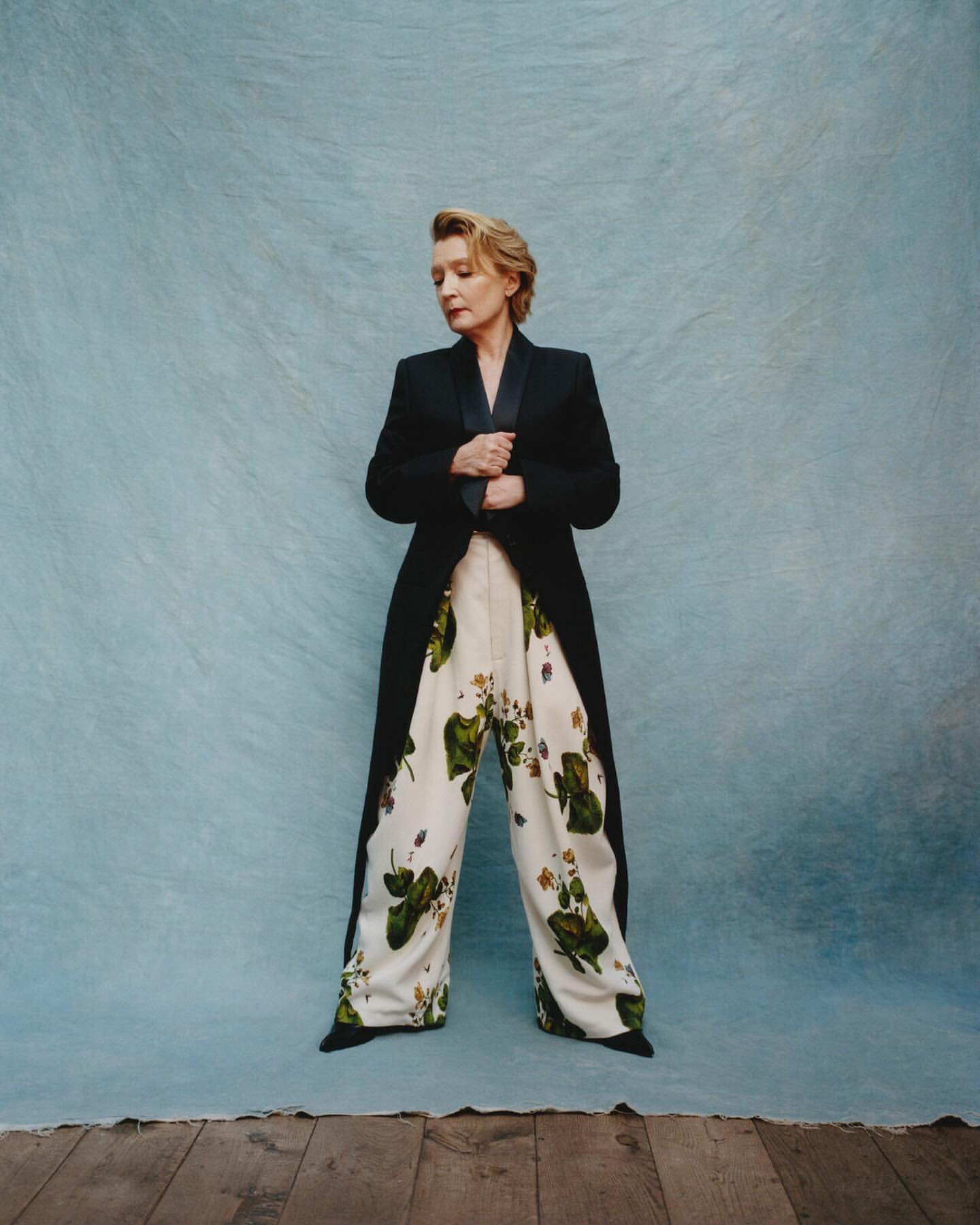 Woman in a long black jacket and floral print pants stands in front of a textured light blue backdrop, posing for a fashion shot.
